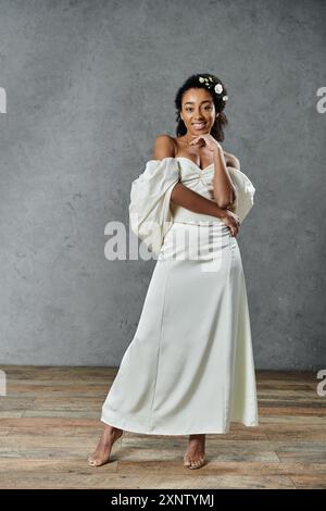 Una splendida sposa afroamericana con un abito da sposa bianco e una corona floreale si posa su uno sfondo grigio. Foto Stock