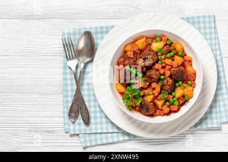 stufato turco di polpette, sulu kofte, piatto abbondante con patate, carote e piselli verdi sospesi in salsa di pomodoro servito in un recipiente con forchetta e cucchiaio su w. Foto Stock