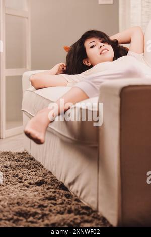 Stile di vita Una giovane donna si rilassa sul suo divano a casa, godendo di una giornata tranquilla per se stessa, incarnando cura di sé e benessere con un sorriso spensierato Foto Stock