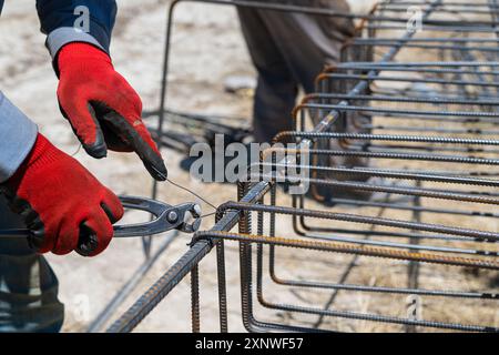 Un operatore utilizza un filo di legatura in acciaio per fissare le barre di acciaio alle barre di rinforzo. Primo piano. Strutture in cemento armato - lavorazione a maglia di un rinforzo in metallo Foto Stock