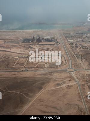 Questa fotografia aerea cattura un'area industriale circondata da un vasto paesaggio desertico in Oman, sottolineando il netto contrasto tra lo sviluppo umano e l'ambiente naturale circostante. L'orizzonte costiero aggiunge un tocco sereno al terreno accidentato. Foto Stock