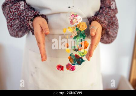 Primo piano di mani femminili ben curate che tengono una tavolozza di plastica con colori misti. Giovane donna in abiti casual e grembiule in studio d'arte. Ragazza alla painti Foto Stock