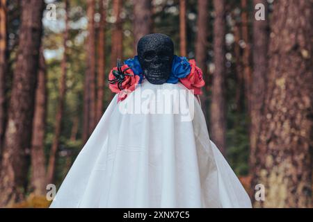 La figura della sposa spettrale drappeggiata di bianco con una maschera cranica nera ornata da fiori blu e rossi si staglia nella foresta, guardando la fotocamera Foto Stock