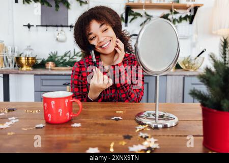 Grazioso africano in pigiama rosso a quadri è seduto a tavola e applica il trucco con un pennello. Ragazza nera in una cucina accogliente con decorazioni natalizie e tazza. H Foto Stock