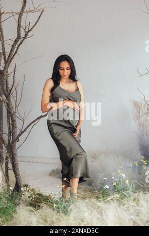 Una giovane donna incinta in un abito di raso verde in piedi nell'erba in estate. Un'affascinante donna incinta si siede di profilo. Una donna serena Foto Stock