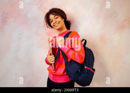 La giovane donna africana in maglione rosso a righe con zaino regge un laptop. Una bella studentessa nera sta andando in classe. Affascinante adolescente riccio in c Foto Stock