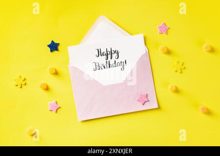Cartolina happy Birthday scritta a mano in busta rosa, top shot su sfondo giallo con stelle glitter e caramelle Foto Stock
