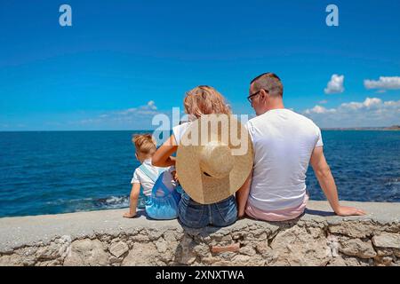 Vista posteriore di padre madre senza volto e figlio piccolo in abbigliamento casual seduto su recinzione rocciosa e guardando mare blu calmo e cielo in vacanza Foto Stock