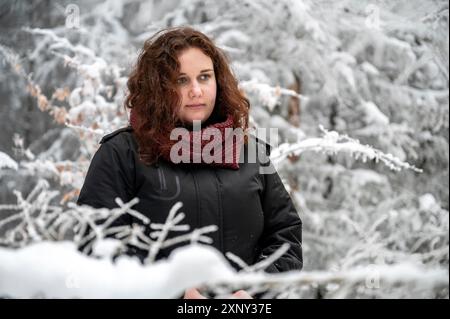 Giovane donna con capelli ricci marroni e premurosa espressione facciale è in piedi in mezzo a una foresta innevata durante l'inverno Foto Stock