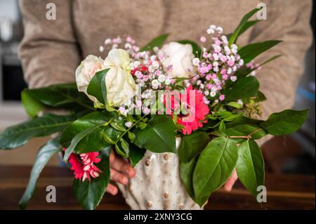 Donna in primo piano con pullover beige e jeans che tiene un bel bouquet di fiori in un vaso, al coperto Foto Stock