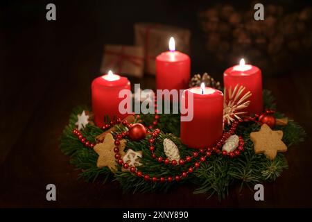 Corona dell'avvento da rami di abete con candele accese di colore rosso e decorazioni natalizie, alcuni regali sfocati sullo sfondo scuro, spazio copia, messa a fuoco selezionata Foto Stock