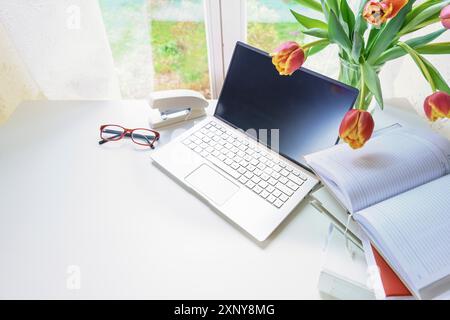 Postazione di lavoro accanto a una finestra con laptop, organiser, raccoglitore ad anelli e un mazzo di tulipani rossi su una scrivania bianca, concetto per lo studio o l'ufficio domestico Foto Stock