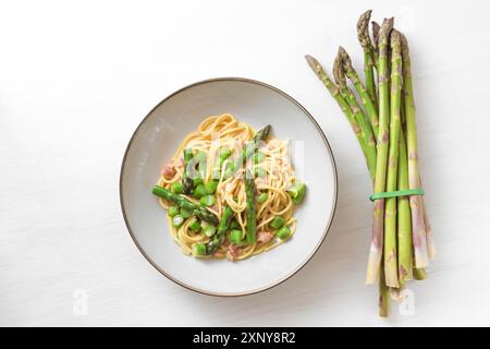 Asparagi verdi con spaghetti, bacon e salsa carbonara su un piatto accanto a un fascio di verdure crude su un tavolo di legno dipinto bianco, copia Foto Stock