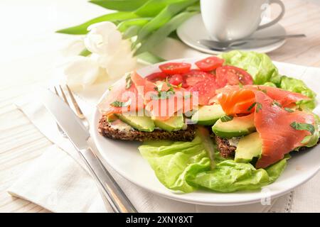 Panino intero con avocado e salmone affumicato con lattuga e insalata di pomodoro, posate e tazza da caffè su un tavolo di legno dipinto di bianco, spazio per le copie Foto Stock