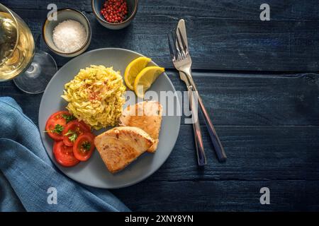 Gustosa portata di pesce con bistecche di tonno fritte, cavolo savoy, pomodori e limone serviti con vino, spezie e posate su un tavolo rustico in legno blu scuro Foto Stock