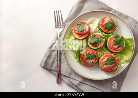 Fette di zucchine al forno con pomodoro, parmigiano e guarnigione di prezzemolo su un piatto con lattuga, tovagliolo grigio su un tavolo dipinto bianco con copia Foto Stock