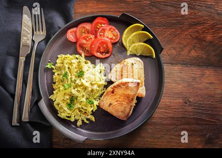 Bistecche di tonno fritte con cavolo sabaudo, limone e pomodori su un piatto scuro e un rustico tavolo in legno con tovagliolo e posate, spazio per copiare, angolo alto Foto Stock