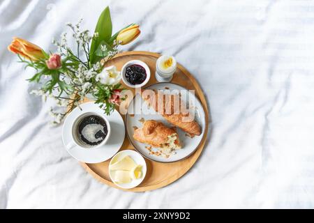 Colazione tranquilla a letto, vassoio di legno con croissant, caffè, marmellata, uova, burro e fiori su biancheria bianca verde grigia, domenica o mattina festiva Foto Stock