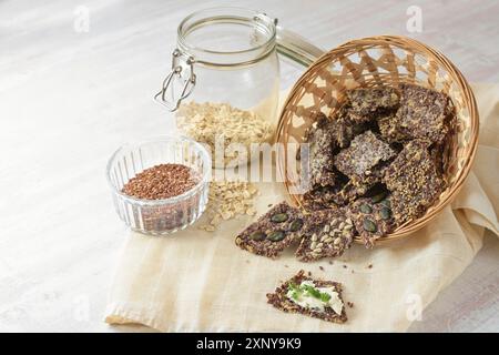 Spuntini croccanti di semi di lino, fiocchi d'avena e semi vari in un cesto, uno con burro e guarnigione di prezzemolo, ingredienti in vasetti di vetro, tavolo leggero Foto Stock
