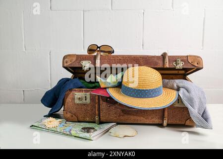 Valigia vintage con accessori da viaggio come cappello di paglia, asciugamani, passaporto, mappa e occhiali da sole per le vacanze al mare in estate, pareti bianche ruvide Foto Stock