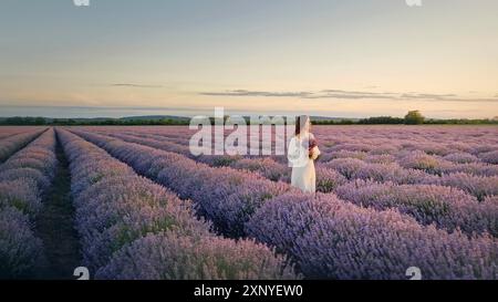 Pittoresca scena estiva con una giovane donna in abito bianco in attesa su un campo di lavanda al tramonto. Splendida sposa con fiori viola di lavandula Foto Stock