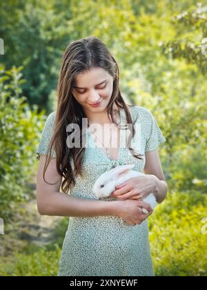 Grazioso ritratto di una giovane donna che tiene un coniglietto bianco tra le braccia su uno sfondo verde. Ragazza che accarezza un adorabile coniglio Foto Stock