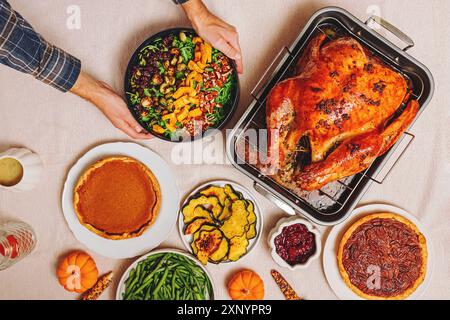 Uomo che mette un'insalata fresca sul tavolo piena di piatti tradizionali del Ringraziamento con tacchino arrosto dorato, fagiolini, zucca arrosto e torta Foto Stock