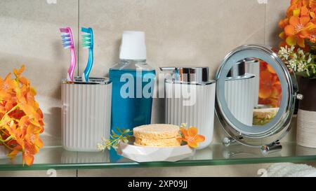 Vista ravvicinata del ripiano in vetro del bagno con bottiglia per il risciacquo vocale, spazzolino da denti, specchio, bottiglia di sapone liquido, spugne per doccia e fiori Foto Stock