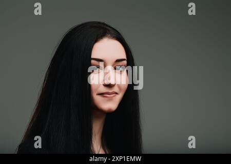 Giovane donna con lunghi capelli scuri nel profondo del pensiero, sorridente delicatamente con un'espressione serena, contemplando decisioni importanti contro un bac grigio neutro Foto Stock