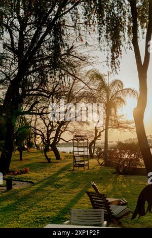 Uno scatto verticale di alberi alti e sedie vuote in un parco sul lago, con una scena di tramonto sullo sfondo Foto Stock