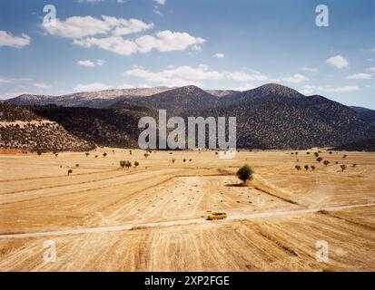 Un taxi giallo viaggia lungo una strada sterrata attraverso un vasto campo dorato di paglia, con montagne sullo sfondo ad Antalya, Turchia. La giornata di sole mette in risalto un senso di viaggio e avventura. Foto Stock