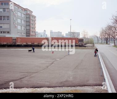 Famiglia con due bambini e un cane che gioca in un parcheggio industriale deserto, con un treno merci sullo sfondo. Una metafora per trovare gioia in luoghi desolati. Foto Stock