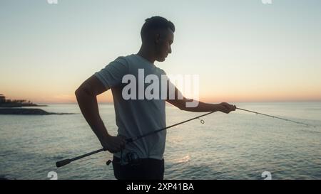 L'uomo con una canna da pesca si erge sulla riva e si gode un bellissimo tramonto. Pescatore che pesca sull'oceano e che ama la natura Foto Stock