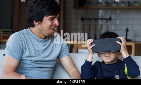 Un padre e un figlio che trascorrono del tempo di qualità a casa. Ragazzo che indossa il visore VR e sperimenta una tecnologia coinvolgente, mentre il padre guarda e sorride. Moderno Foto Stock