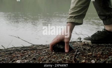 Un primo piano della mano di una persona che prende una pietra sul lungolago. Le calme acque del lago e lo sfondo nebbioso suggeriscono un ambiente all'aperto sereno e tranquillo Foto Stock