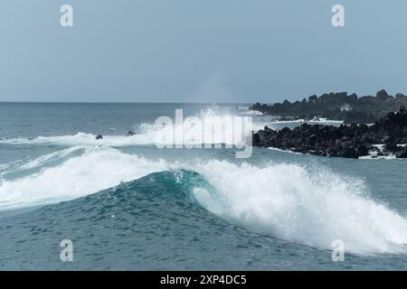Potenti onde oceaniche si scontrano contro rocce vulcaniche nere sotto un cielo blu Foto Stock