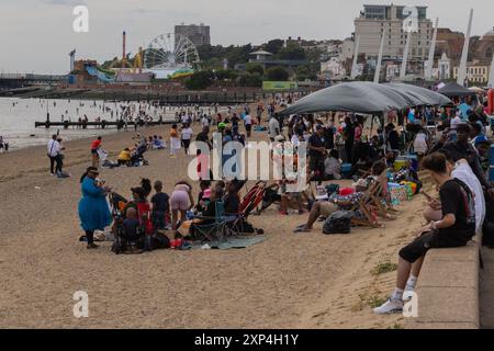 Southend on Sea, Regno Unito. 3 agosto 2024. Una vivace spiaggia che si riunisce con numerose persone sedute sotto ombrelloni e su sedie pieghevoli, godendosi del tempo libero vicino alla costa. Una ruota panoramica e giostre sono visibili sullo sfondo, suggerendo la vicinanza a un'area di intrattenimento sul mare. I groynes di legno si estendono fino al mare e gli edifici a più piani fiancheggiano la costa, indicando un ambiente urbano. La polizia dell'Essex ha implementato un ordine di dispersione e i poteri della sezione 60 per 48 ore a causa di precedenti violenti disordini lungo il lungomare e High Street. Penelope Barritt/Alamy Live News Foto Stock