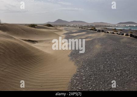 La strada sulla spiaggia di Famara, invasa dalle dune di sabbia. La Caleta de Famara sullo sfondo. Lanzarote, isole Canarie Foto Stock