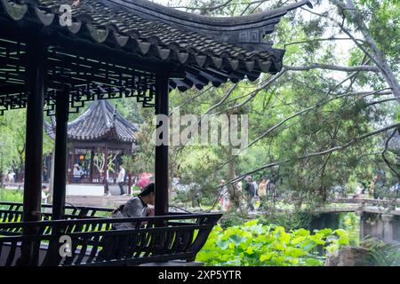 Suzhou, Cina - 11 giugno 2024: Una donna siede in un tradizionale padiglione cinese che si affaccia su un tranquillo laghetto con foglie di loto, un ponte e un verde lussureggiante Foto Stock