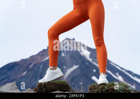 Le gambe atletiche della donna fanno esercizio fisico in montagna con il vulcano sullo sfondo. Primo piano di gambe femminili in una figura arancione che avvolge i leggings sportivi Foto Stock