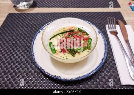 Uovo in camicia con purè di patate, asparagi e prosciutto in una ciotola di vetro trasparente su un piatto bianco su un tovagliolo in tavola in un ristorante Foto Stock