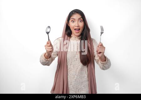 Scioccata donna musulmana asiatica in velo hijab che tiene posate su entrambe le mani e si sente sorpresa, in piedi su sfondo bianco isolato Foto Stock