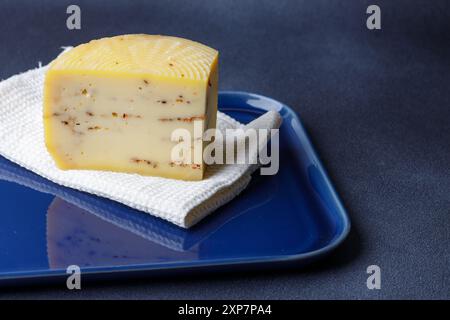 Un blocchetto di formaggio artigianale infuso di erbe si appoggia su un panno bianco, posto su un piatto di ceramica blu, evidenziandone le ricche consistenze e il c Foto Stock