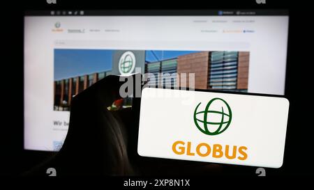 Persona che possiede uno smartphone con logo della società di vendita al dettaglio tedesca Globus Holding GmbH Co. KG davanti al sito web. Mettere a fuoco il display del telefono. Foto Stock