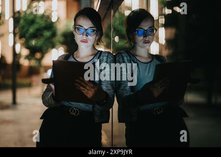 Donna d'affari che lavora su un tablet di notte, riflessa su una finestra di vetro con luci stradali sullo sfondo Foto Stock