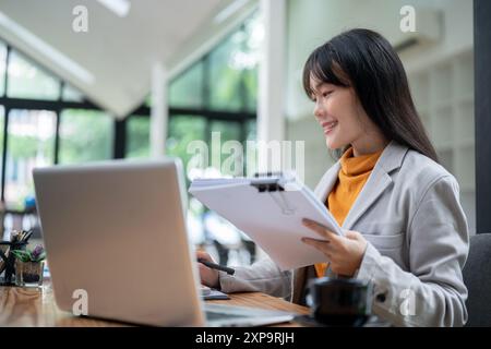 Una giovane donna d'affari asiatica, bella e soddisfatta, sta lavorando nel suo ufficio, esaminando i rapporti aziendali e gestendo i documenti aziendali alla sua scrivania. Foto Stock