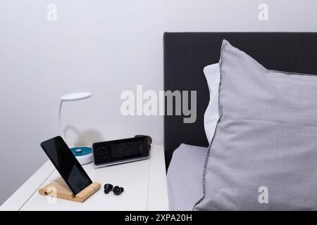 Smartphone sul comodino con auricolare, lampada, orologio e letto come sfondo Foto Stock