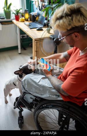 L'uomo disabile in sedia a rotelle guarda le previsioni meteo sullo schermo del telefono, siede accanto al ventilatore con il cane a casa. Foto Stock