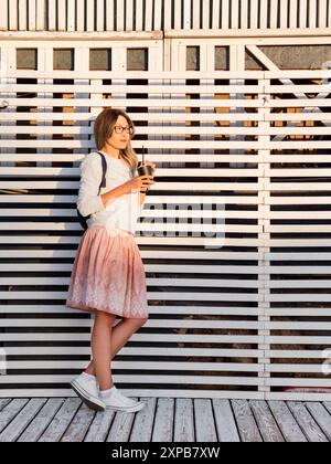 Donna con una tazza di caffè incontra il tramonto sul molo di legno. La donna con i capelli ricci sta godendo la natura all'aperto. Abbigliamento casual estivo. Vibrazioni estive. Foto Stock