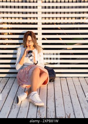 Donna con una tazza di caffè incontra il tramonto sul molo di legno. La donna con i capelli ricci sta godendo la natura all'aperto. Abbigliamento casual estivo. Vibrazioni estive. Foto Stock